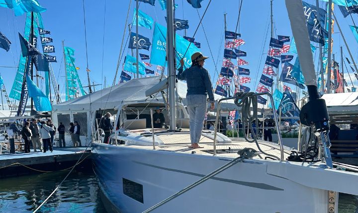 A woman stands on the deck of a wrapped Beneteau sailboat at a boat show, showcasing the premium boat wrap by AP Wraps amid a vibrant display of flags and attendees.
