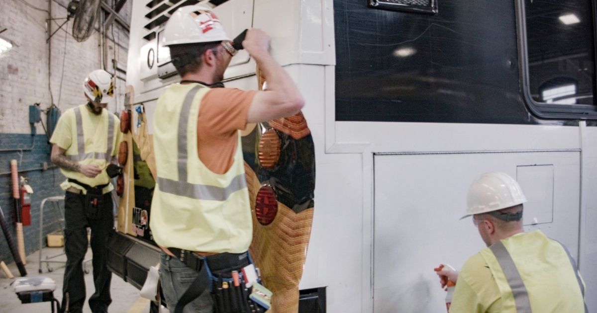 baltimore-transit-bus-wrap-graphics-installation-osha-gear Technicians in OSHA-compliant PPE installing vinyl bus wrap graphics on a Baltimore Link transit bus inside a service facility